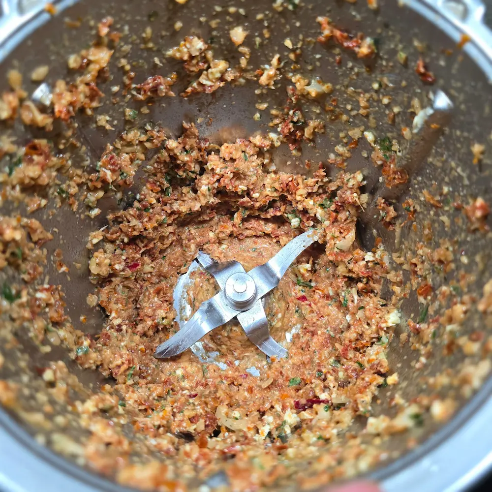 Rozdrobnione suszone pomidory, cebula, czosnek i zioła na dnie naczynia miksującego Thermomix