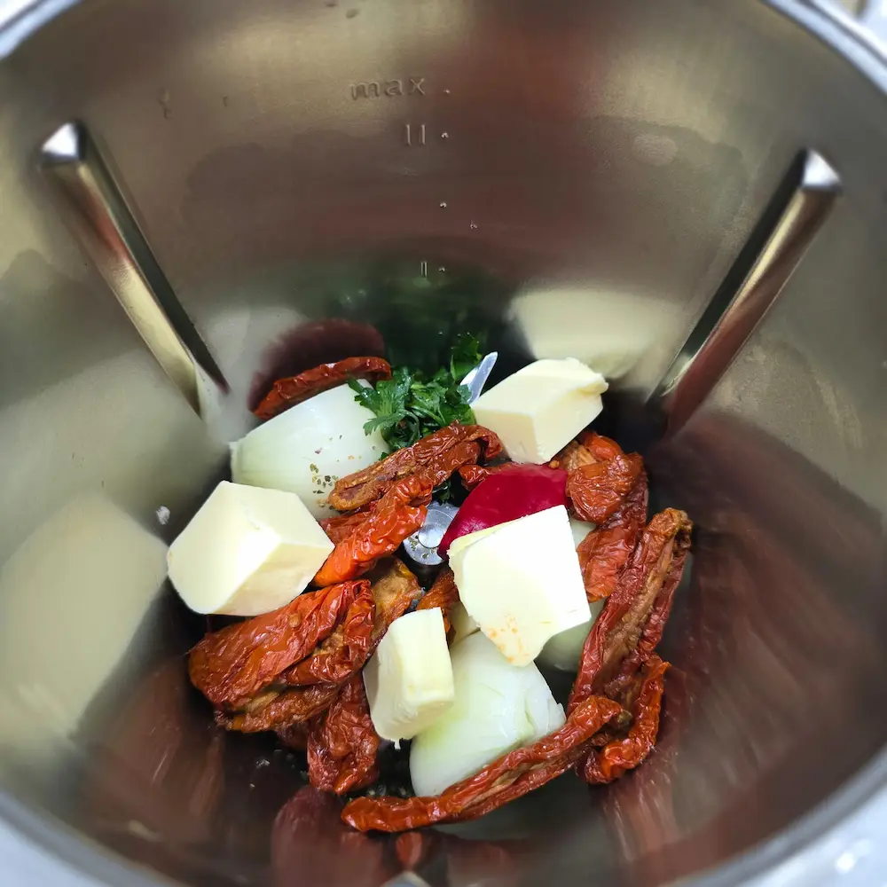 Suszone pomidory, kawałki cebuli, masło i świeże zioła w naczyniu miksującym Thermomix przed rozdrobnieniem